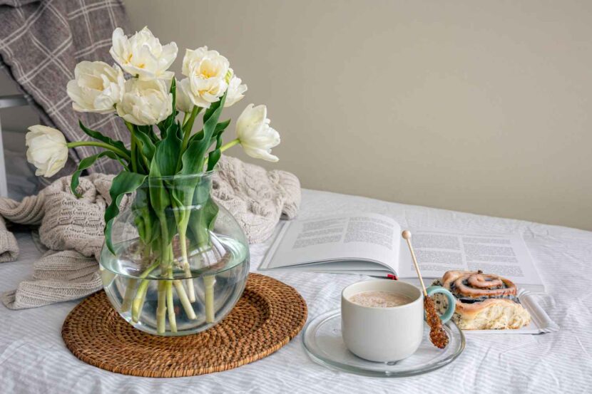 A bright and airy aesthetic morning scene with sunlight streaming through an open window onto white linen sheets and a small vase of spring flowers.