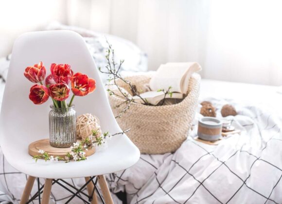 A bright, sunlit bedroom with white linen sheets, flower-shaped pillows, and a small vase of tulips on a wooden nightstand.