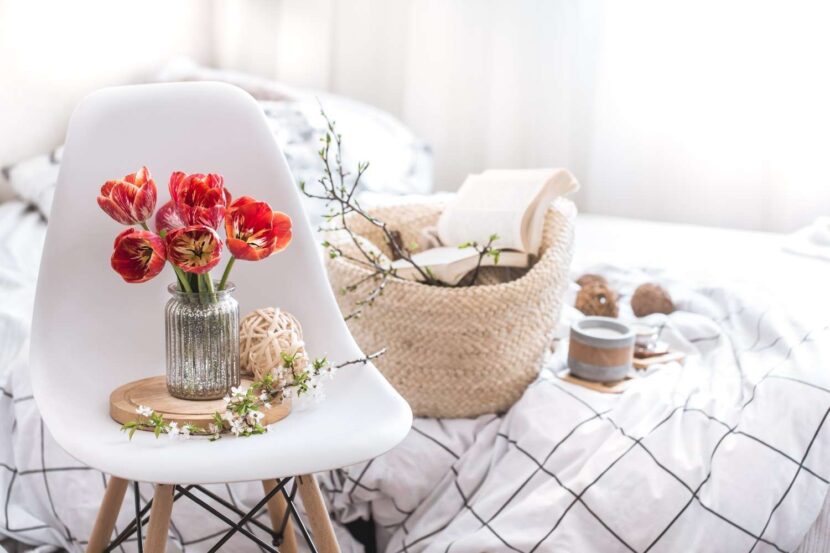 A bright, sunlit bedroom with white linen sheets, flower-shaped pillows, and a small vase of tulips on a wooden nightstand.