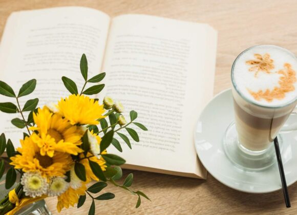 Aesthetic sunny summer desk setup featuring an open journal, a cup of coffee, and a bouquet of yellow flowers in the June sunlight.